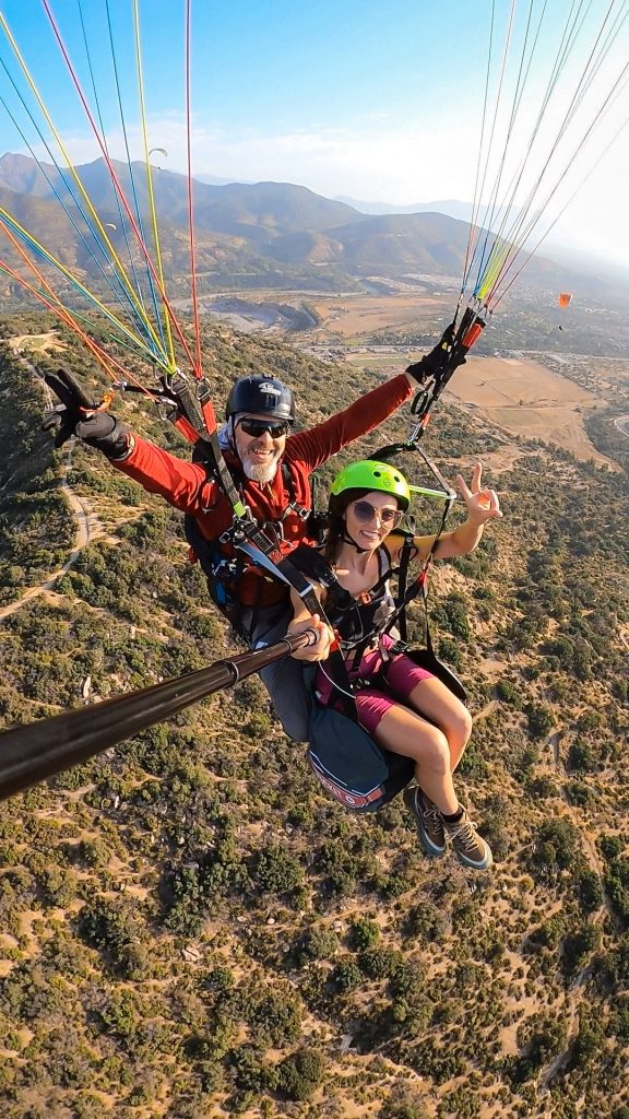 Woman smiling and making a peace sign high above the ground in Chile while paragliding with an expert guide