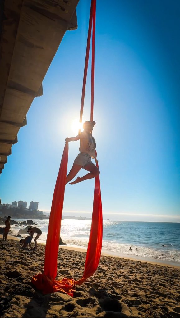 Woman doing aerial silks on the beach in Chile with the sun and ocean behind her