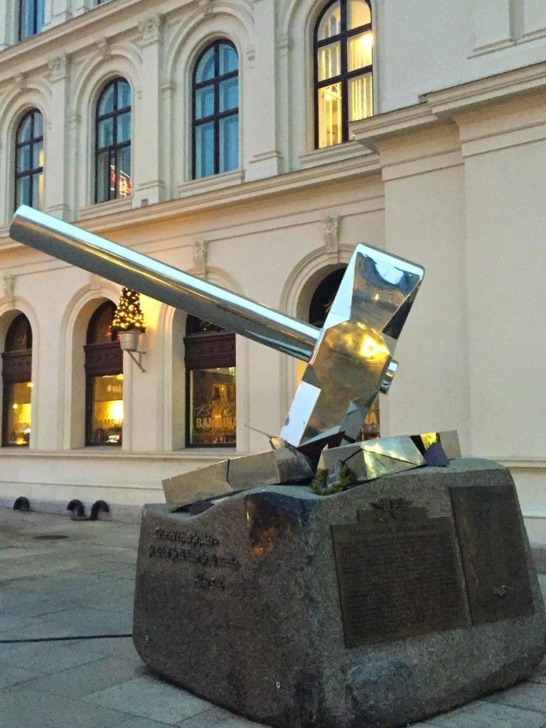 Knus Nazismen shiny silver hammer statue, striking and breaking a sheet of metal on top of a large stone. Statue is in front of a beautiful building with Christmas lights in Oslo, Norway in winter