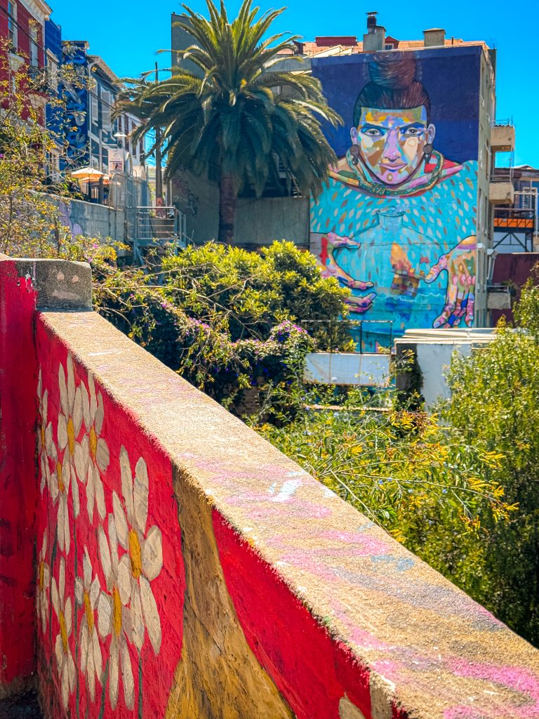 Huge mural of a woman on the side of a building overtop of a city wall painted with flowers 