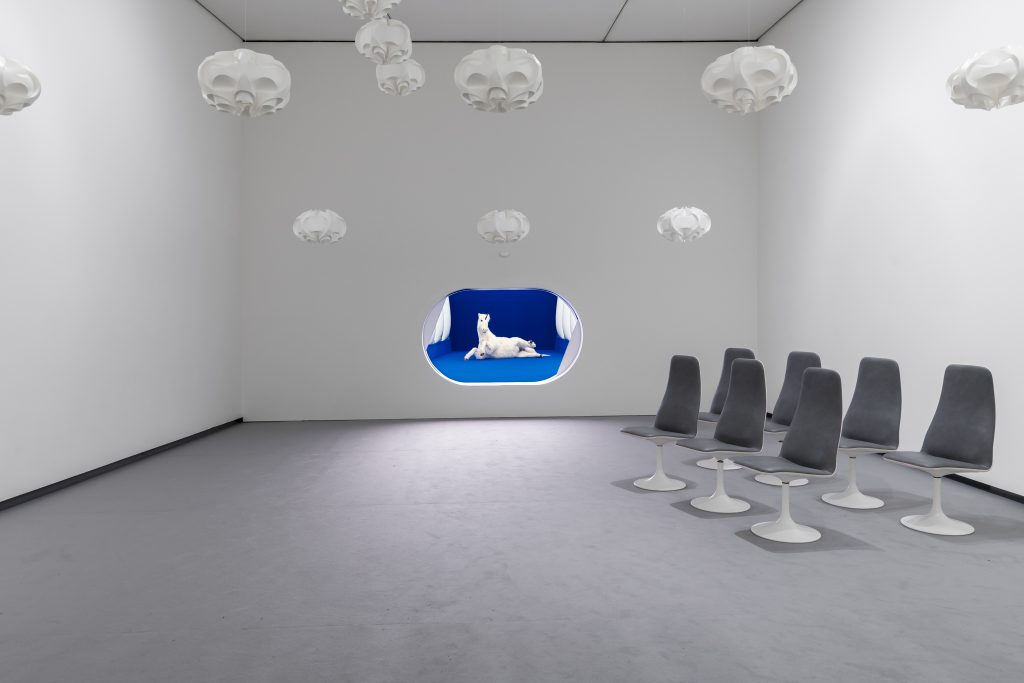 White room with clouds hanging from the ceiling with a blue cut out in the wall holding a white horse