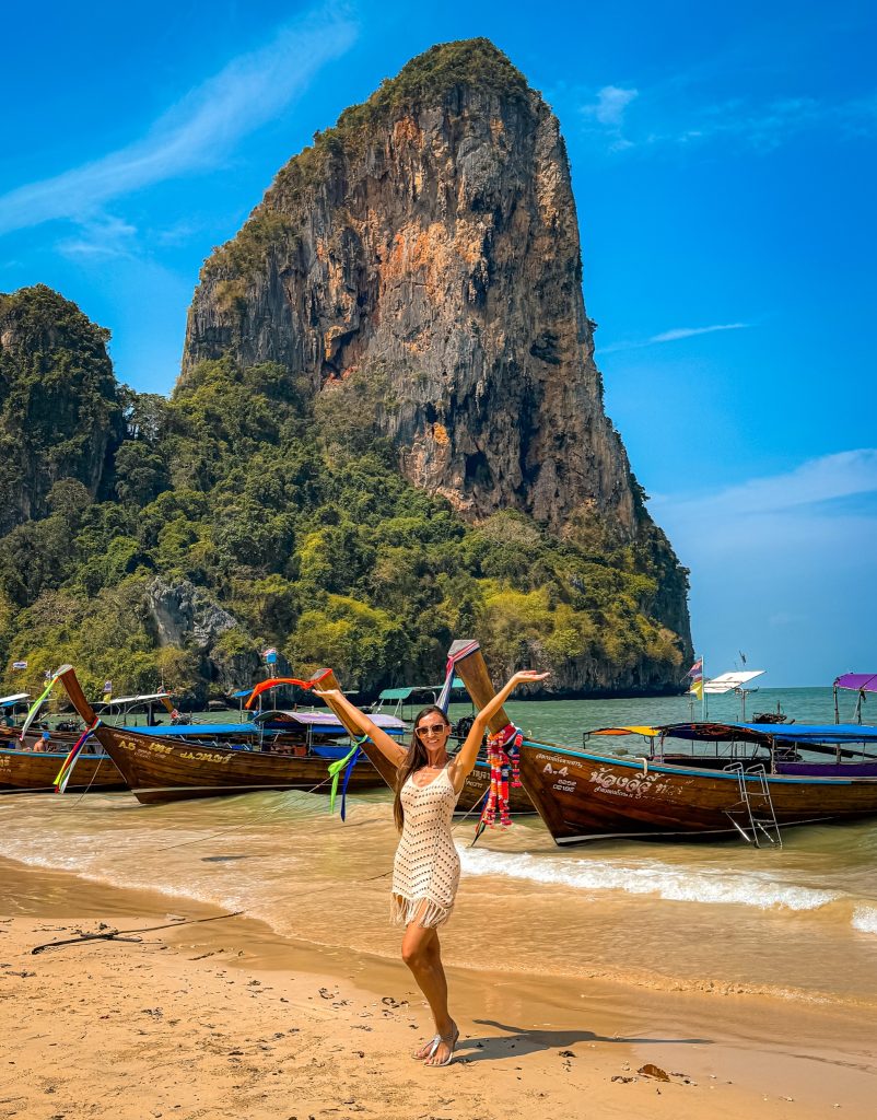 Woman posing with raised arms beside colorful longtail boats on Railay Beach.