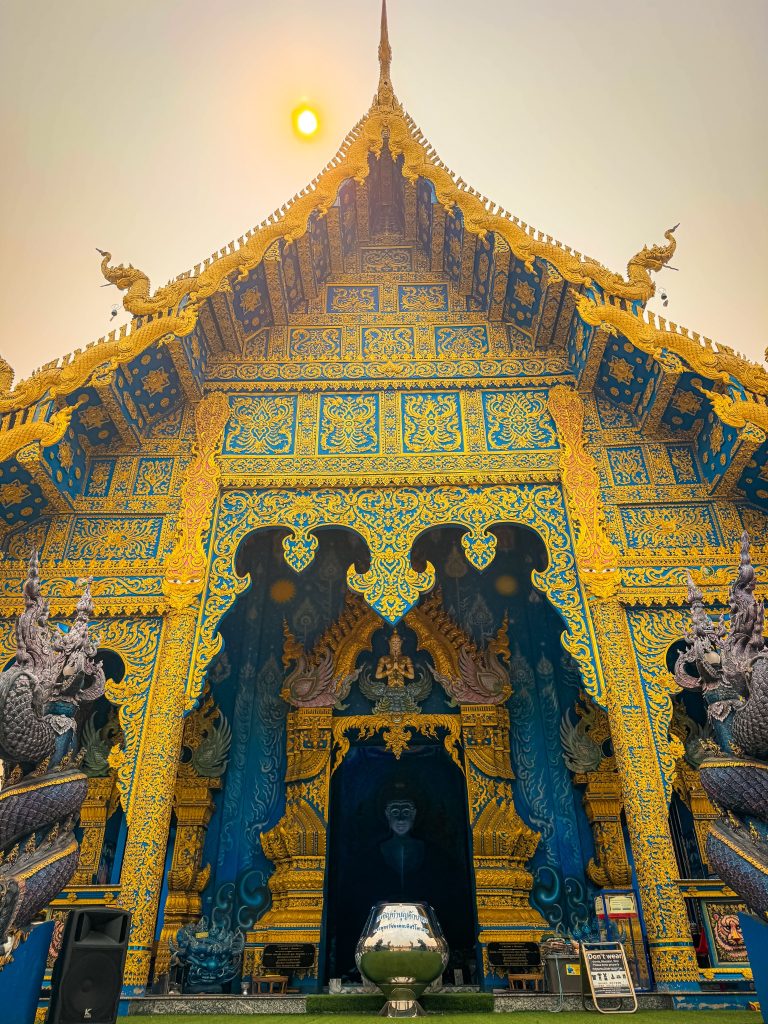 Front facade of the Blue Temple in Chiang Rai with ornate gold details and the sun above the roof.