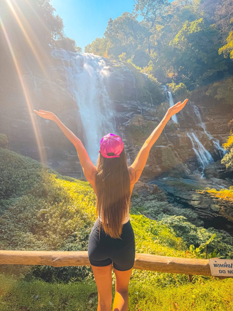 Traveler standing with arms raised in front of Wachirathan Waterfall in Doi Inthanon National Park, Thailand.
