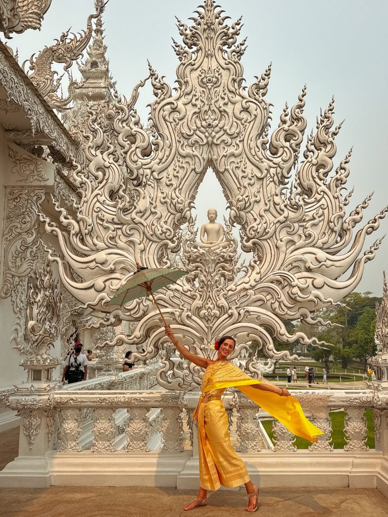 Woman in a gold traditional Thai dress holding a green parasol in front of a white Buddha sculpture at the White Temple.