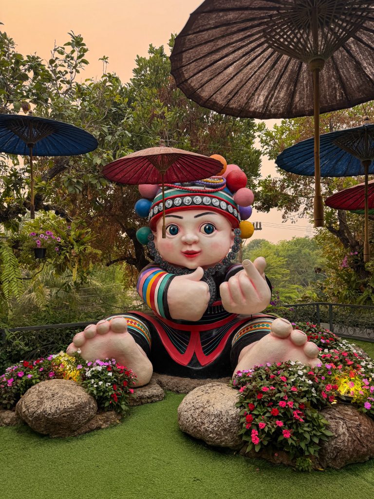 Large colorful hill tribe-inspired statue surrounded by flowers and umbrellas at Lalitta Cafe Chiang Rai.