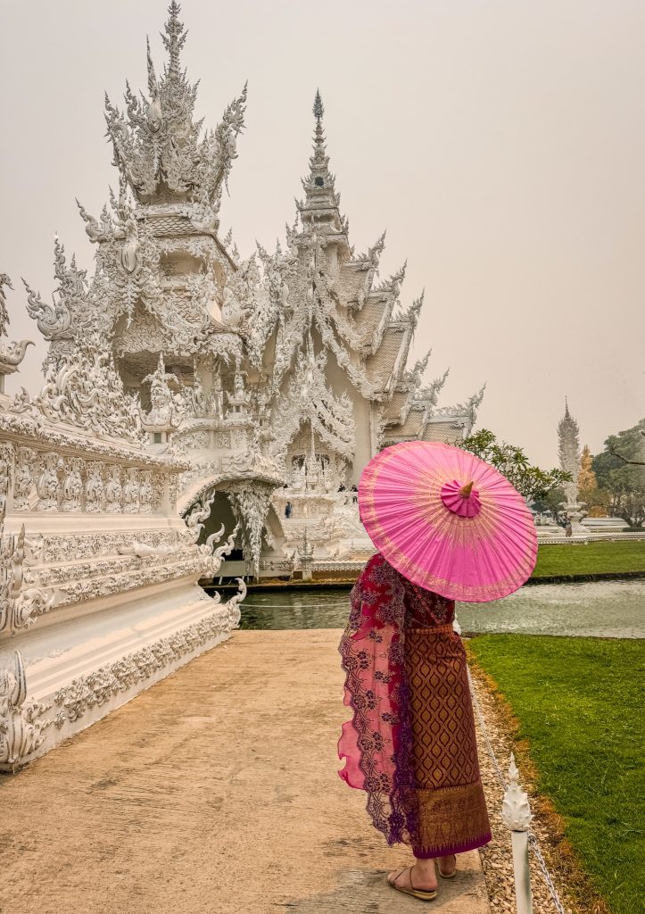 Woman in a burgundy traditional Thai dress carrying a pink parasol beside the White Temple in Chiang Rai.