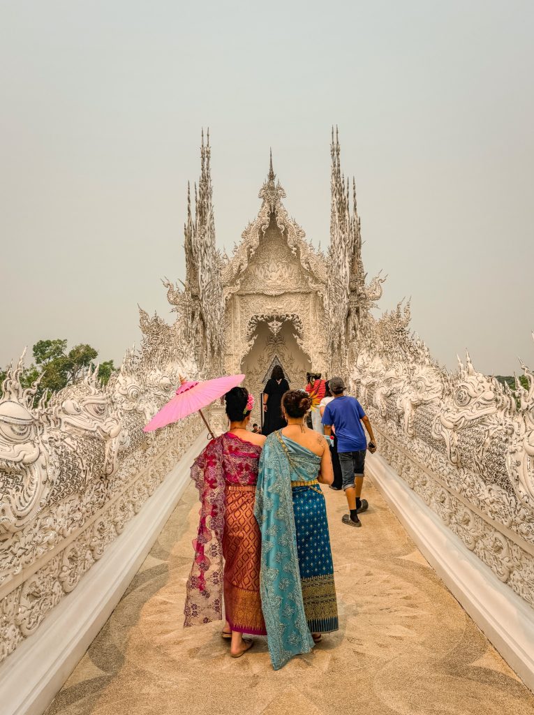 Two women in colorful traditional Thai dresses walking across the ornate bridge toward the White Temple.