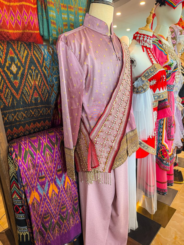 Lavender traditional Thai outfit displayed beside colorful woven textiles at a Chiang Rai dress shop.