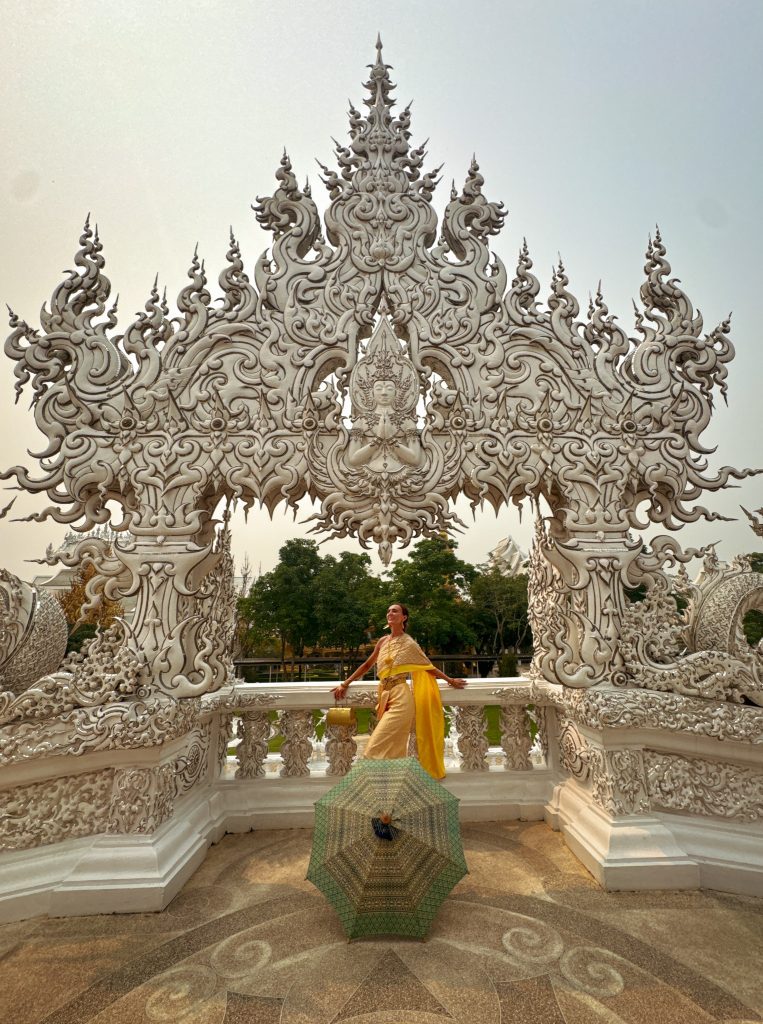 Woman in a gold traditional Thai dress posing beneath an intricately carved arch at the White Temple in Chiang Rai.