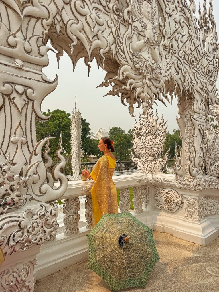 Woman in a gold traditional Thai dress standing among ornate white carvings at the White Temple in Chiang Rai.
