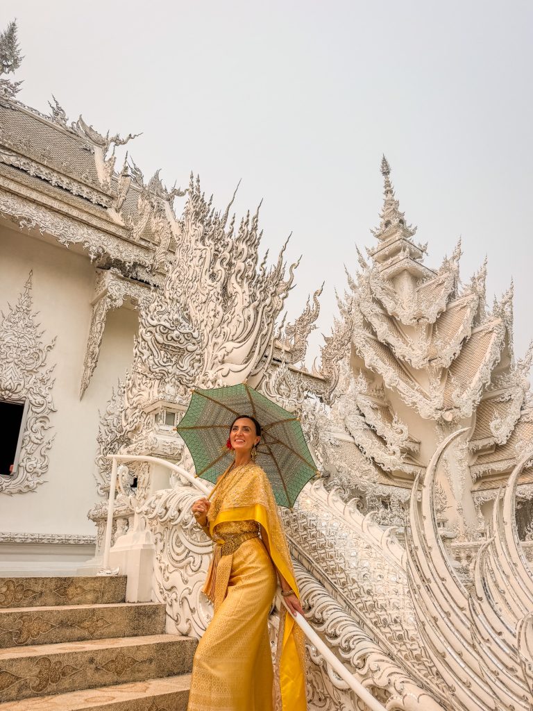 Woman in a gold traditional Thai dress posing beside the detailed white stairway at the White Temple in Chiang Rai.