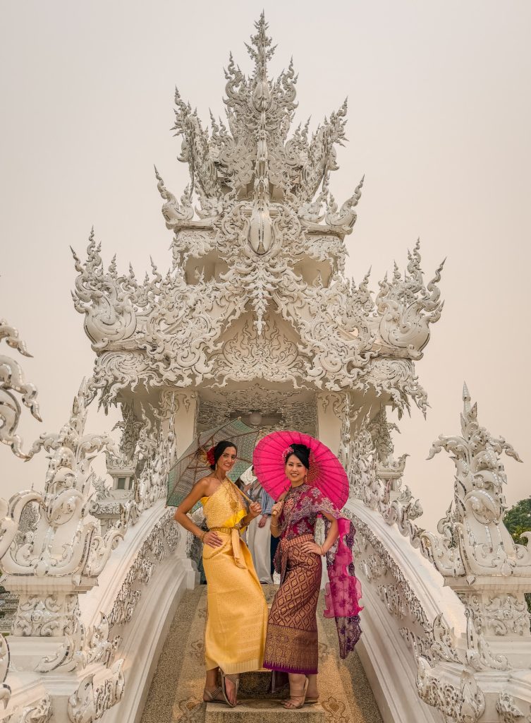 Two women in traditional Thai dresses posing with green and pink parasols on the White Temple bridge.