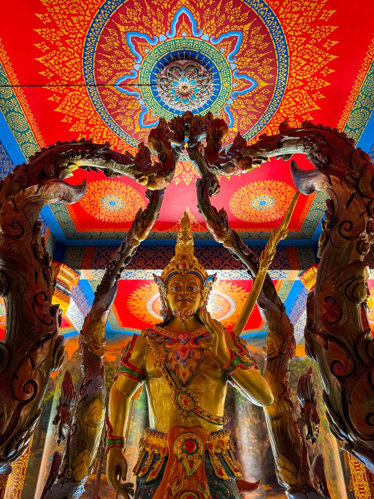 Golden statue with bright, ornate painted ceiling behind it in Thai temple