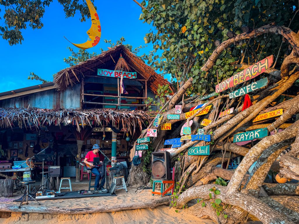 Colorful signs, tree roots, and a musician outside Joy Thai Food on the beach in Krabi.