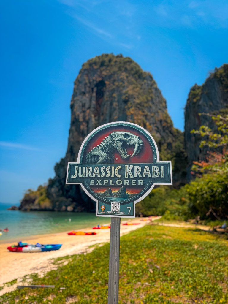 Jurassic Krabi Explorer checkpoint sign with limestone cliffs and kayaks at Phra Nang Beach.