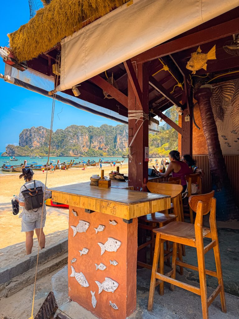 Open-air beachfront cafe seating overlooking longtail boats and limestone cliffs at Railay Beach.