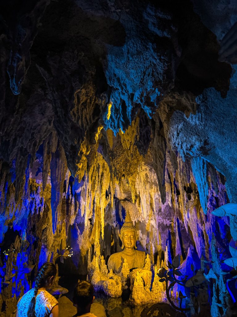 Golden Buddha statue surrounded by stalactites and colorful lighting inside a cave in Chiang Rai.