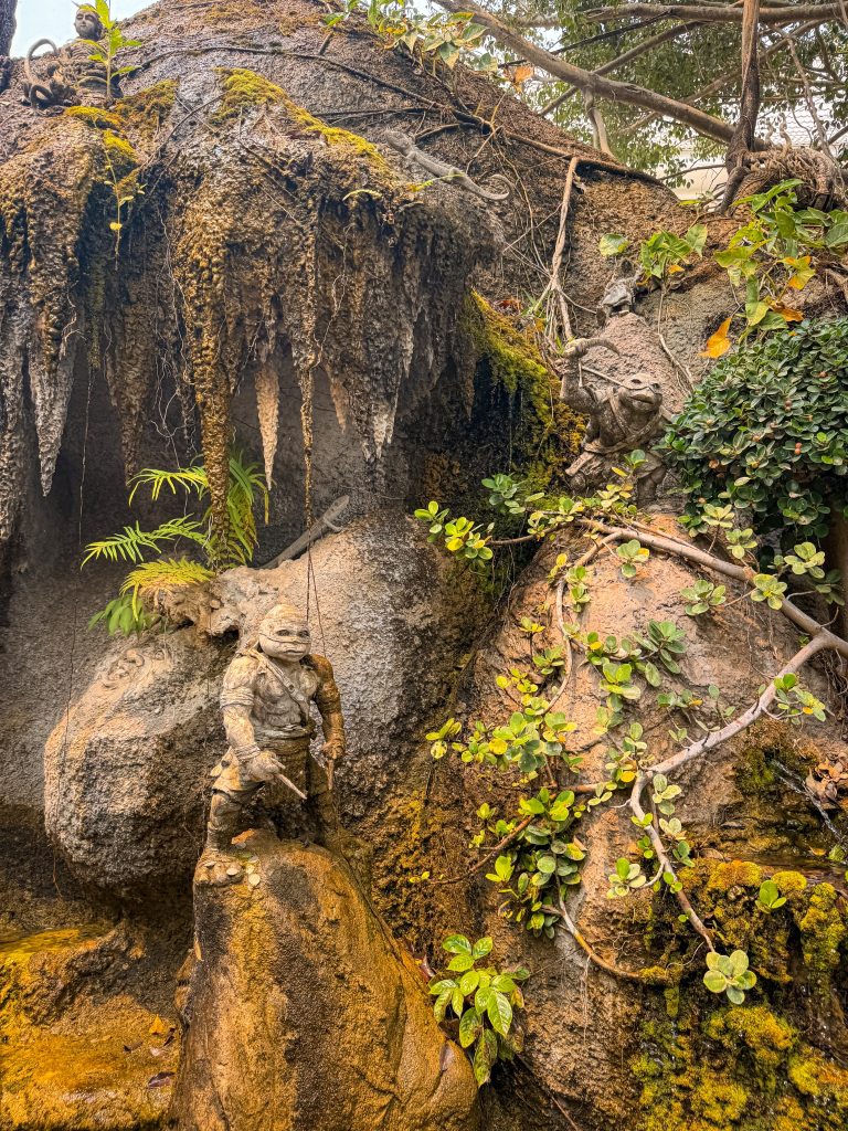 Fantasy character sculptures and moss-covered rock formations near a cave at the White Temple complex.