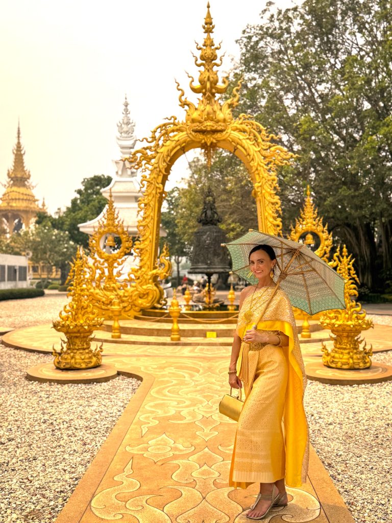 Woman in a gold traditional Thai dress holding a green parasol in front of an ornate golden sculpture.