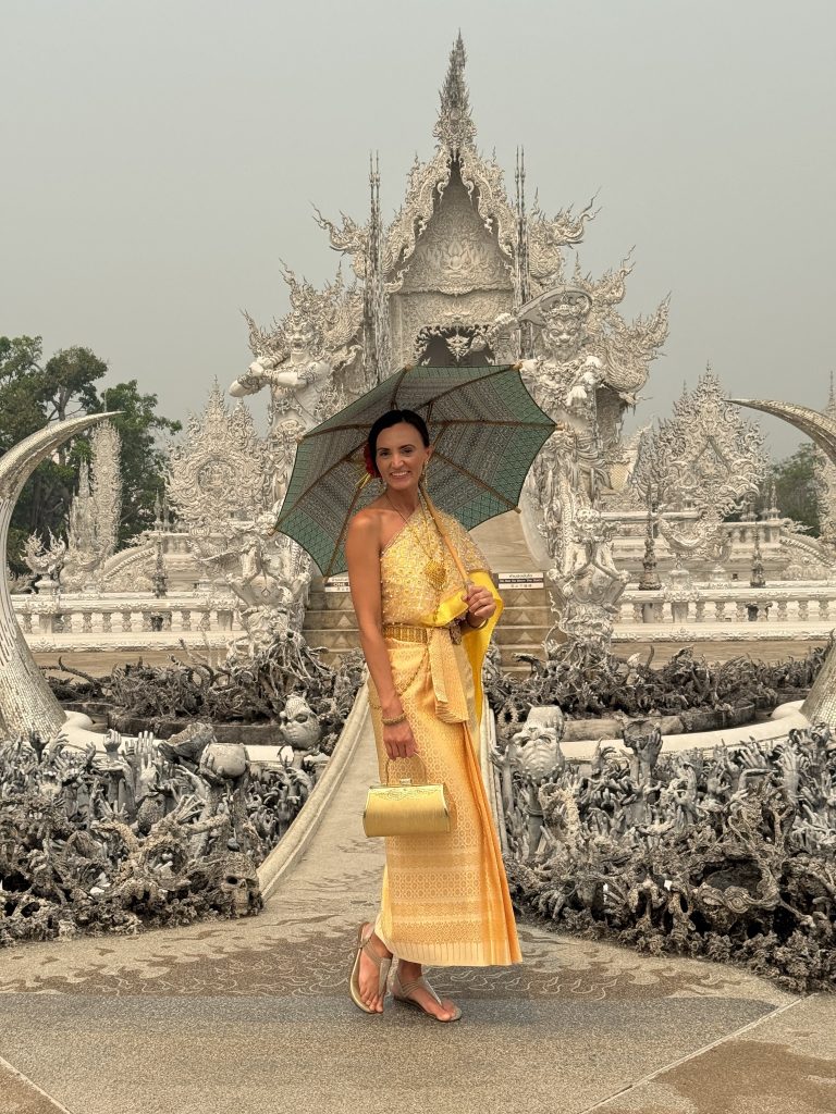 Woman in a gold traditional Thai dress holding a green parasol beside the reaching-hand sculptures at the White Temple.