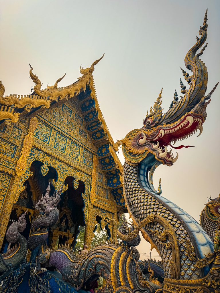 Large blue and gold naga statue beside the ornate exterior of the Blue Temple in Chiang Rai.
