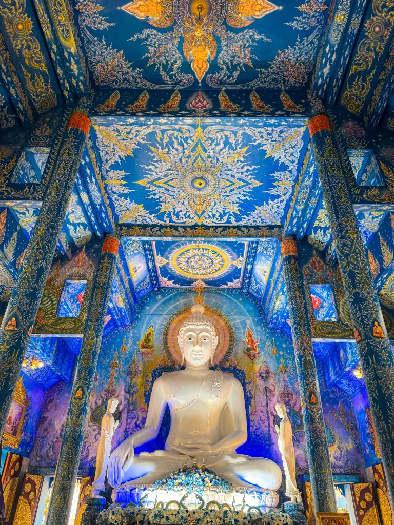 Large white Buddha statue beneath vivid blue and gold murals inside the Blue Temple in Chiang Rai.