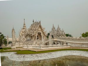 Wide view of the White Temple and its ornate bridge across a reflective pond in Chiang Rai.
