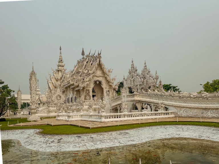 Wide view of the White Temple and its ornate bridge across a reflective pond in Chiang Rai.