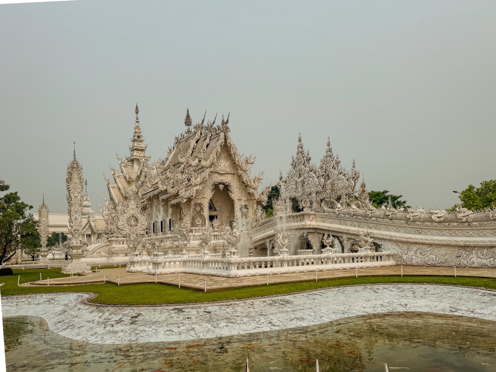 Wide view of the White Temple and its ornate bridge across a reflective pond in Chiang Rai.